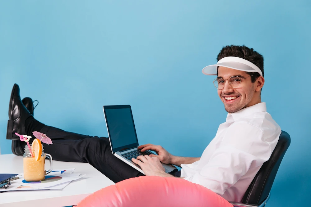 Hombre joven trabajando relajado en un escritorio con un notebook, sonriendo a la cámara y con un visor blanco, mientras descansa los pies sobre la mesa con un trago veraniego al lado.